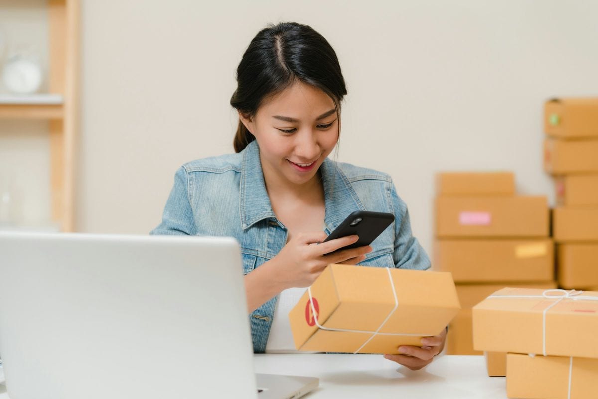 A store owner checking an order on her phone, holding a parcel among shipping boxes: shipping, packing and returns all come out of the margin before the ads get paid